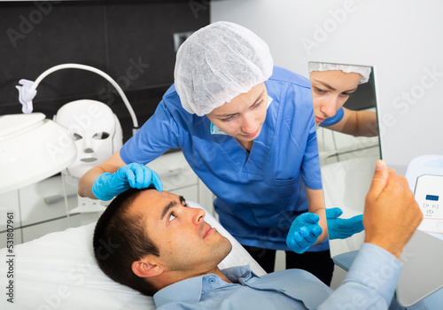 Skilled woman beautician examining face skin of young male patient before cosmetology procedure in beauty clinic