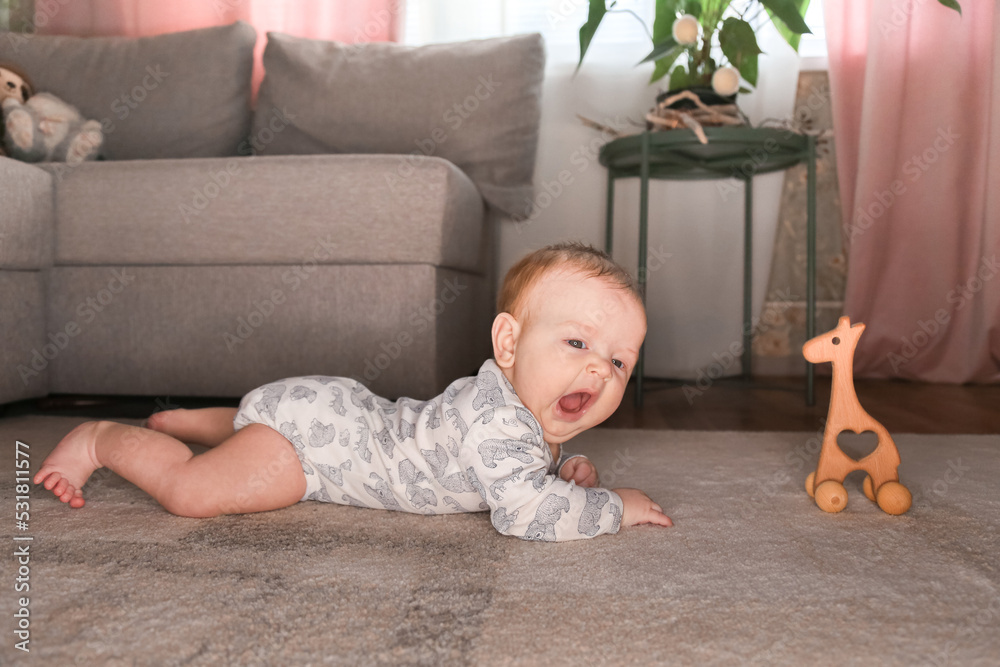Naklejka premium a small child, a baby in a bodysuit lies in the living room on the floor, on the carpet, plays with wooden toys, lifestyle, childhood, baby care, eco-friendly toys, early development