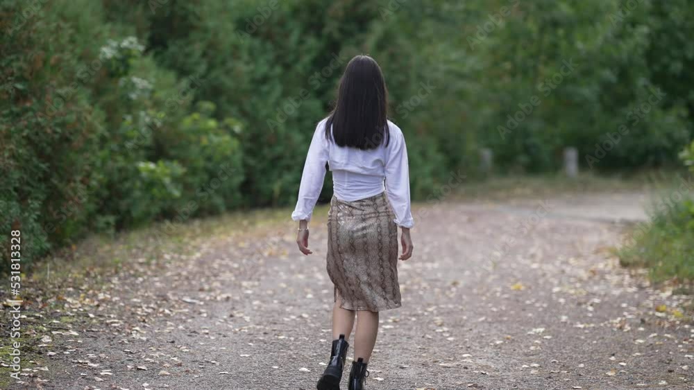 Back view slim elegant young woman walking in slow motion on alley in green park raising hands. Happy confident Asian lady strolling enjoying leisure admiring nature outdoors. Style and femininity Back view slim elegant young woman walking in slow motion on alley in green park raising hands. Happy confident Asian lady strolling enjoying leisure admiring nature outdoors. Style and femininity
