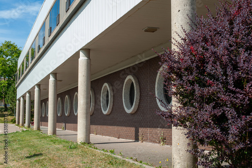 12 large round windows on a brown brick wall in a straight row. The large windows have a grey concrete trim around the exterior glass. There's a walkway along the exterior of the brick building. 