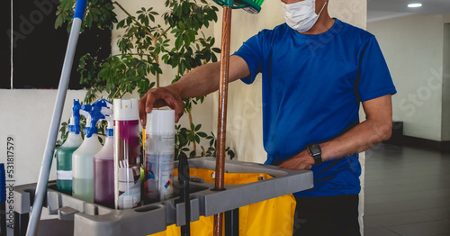 Young latino janitorial worker in blue uniform and trolley with tools and cleaning supplies (sprayers, broom, cleaners, etc) photo