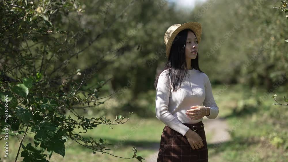 Young attractive slim Asian woman in straw hat looking away standing in sunny park. Portrait of thoughtful calm beautiful lady enjoying summer day outdoors. Slow motion Young attractive slim Asian woman in straw hat looking away standing in sunny park. Portrait of thoughtful calm beautiful lady enjoying summer day outdoors. Slow motion
