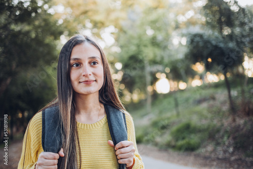 Fototapeta Naklejka Na Ścianę i Meble -  Smiling female university student wearing a yellow sweater and a backpack in the campus park road. Innocent smile