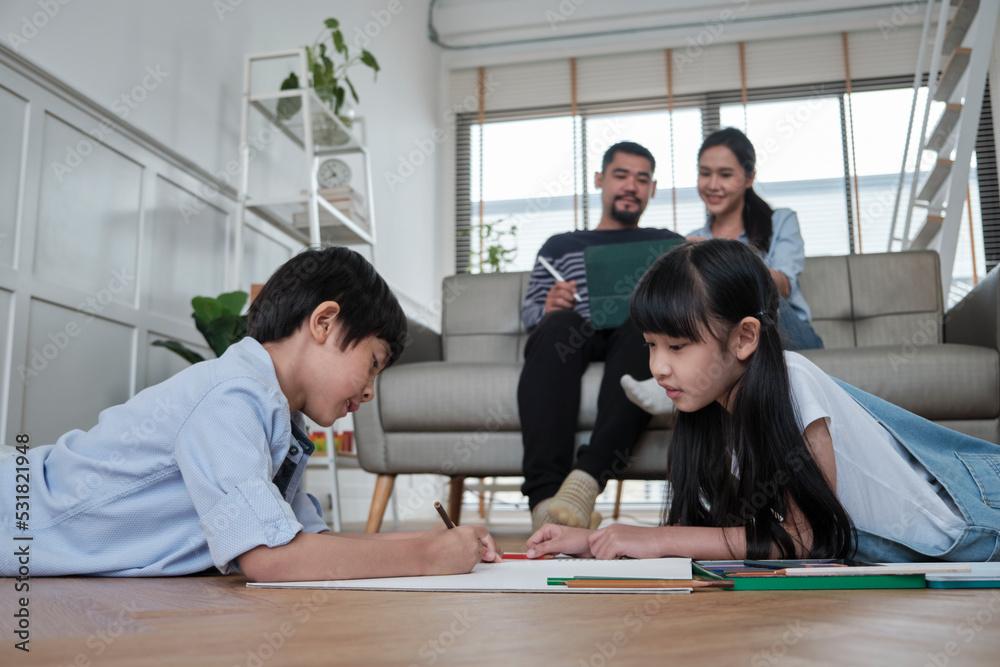 Fototapeta premium Asian Thai siblings are lying on living room floor, drawing homework with colored pencils together, parents leisurely relax on a sofa, lovely happy weekend activity, and domestic wellbeing lifestyle.
