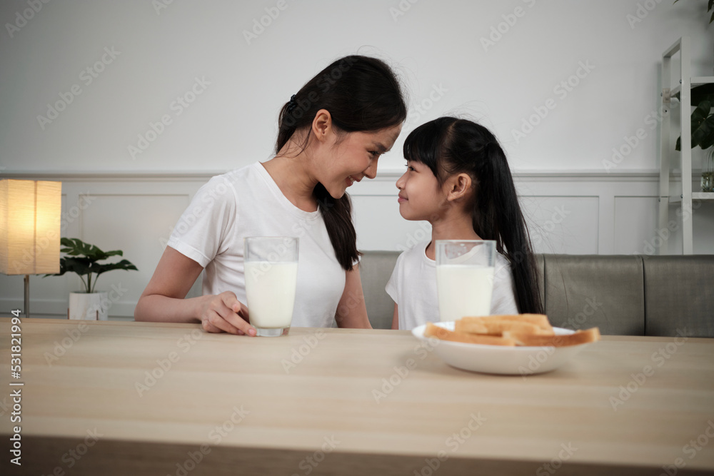 Healthy Asian Thai family, happy little daughter, and young mother face to face at dining table with fresh milk in glass and bread care together, wellness nutrition breakfast meal, morning lifestyle. Healthy Asian Thai family, happy little daughter, and young mother face to face at dining table with fresh milk in glass and bread care together, wellness nutrition breakfast meal, morning lifestyle.