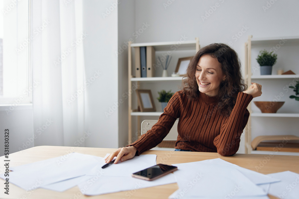 Cheerful happy smiling curly woman writing script counts taxes calculates budget for purchases looks at papers near phone on table. Money Financial literacy concept. Good offer for mortgage credit Cheerful happy smiling curly woman writing script counts taxes calculates budget for purchases looks at papers near phone on table. Money Financial literacy concept. Good offer for mortgage credit