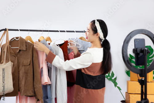 Young asian woman choosing clothes in front of smartphone camera to online selling at home. Live Streaming concept.