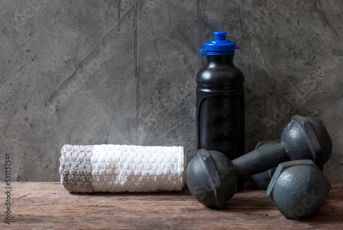 A pair of vintage iron dumbbells with a water bottle and towel on a rustic wooden floor and a cement wall.