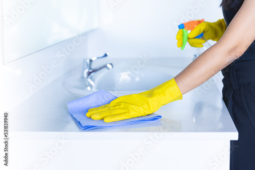 closeup women hand maid or charwoman cleaning modern new basin in bathroom. Water tap clean using yellow gloves and blue cloth with foggy sanitizer spray.