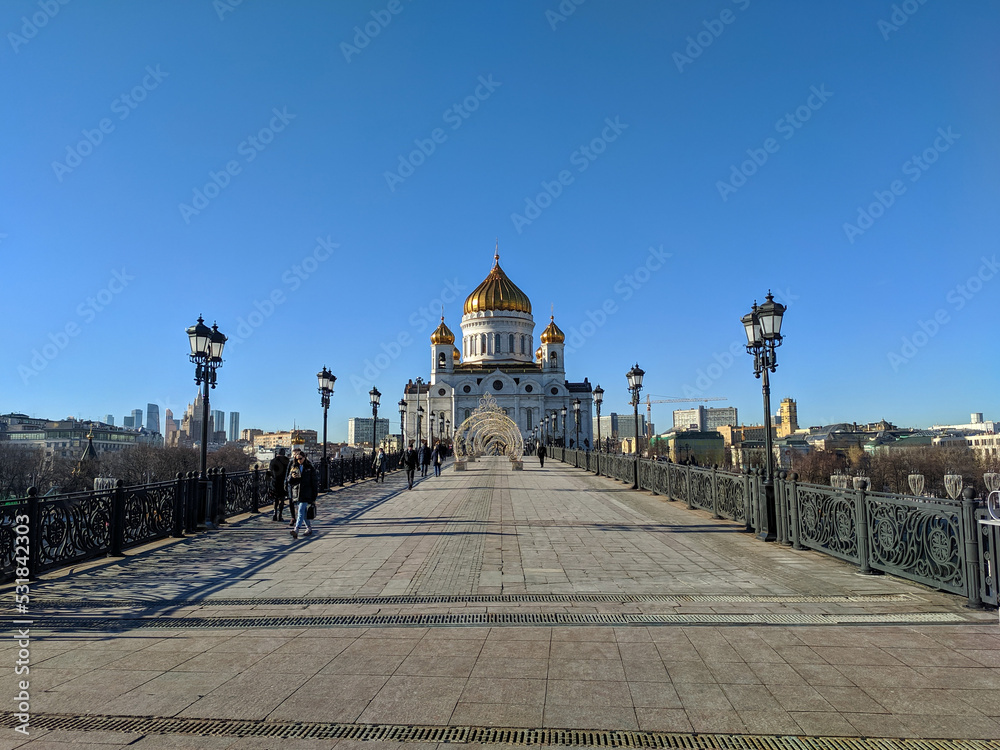 Fototapeta premium Moscow, Russia - March 01, 2022: View of the Cathedral of Christ the Savior from the Patriarchal pedestrian bridge against the blue sky. People are walking on the bridge