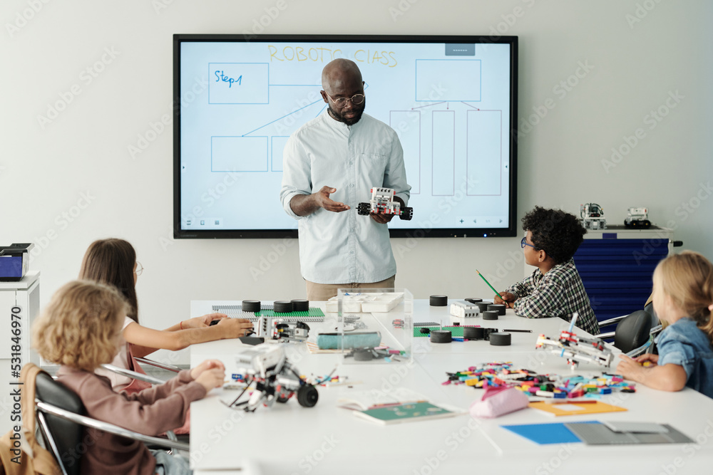 Confident male teacher of robotics explaining intercultural elementary school learners how to connect parts of robot at lesson Confident male teacher of robotics explaining intercultural elementary school learners how to connect parts of robot at lesson