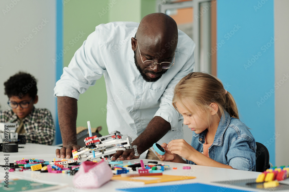 Young teacher of robotics bending over youthful schoolgirl constructing robot while choosing details and connecting them at lesson