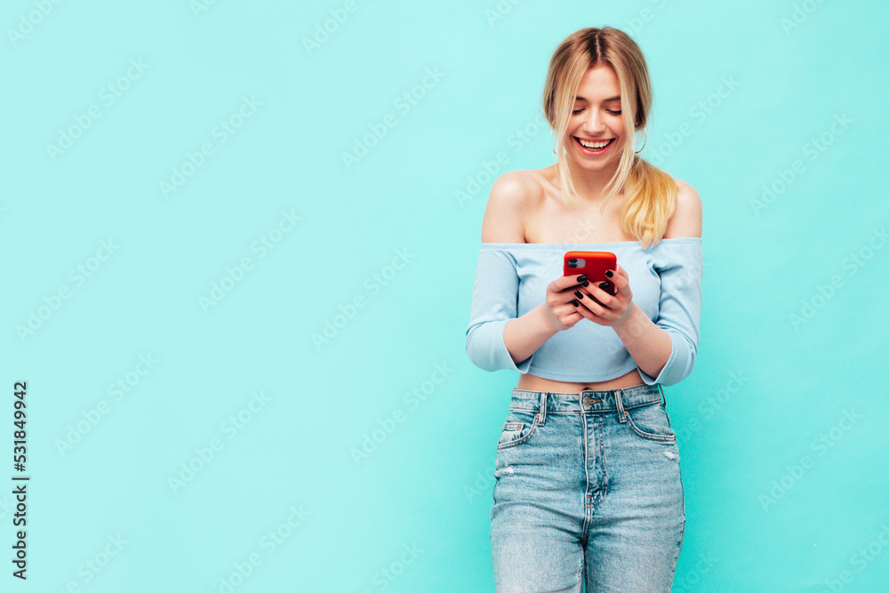 Young beautiful smiling female in trendy summer jeans. Sexy carefree woman posing near blue wall in studio. Positive brunette model looking at cellphone screen. Holding smartphone and using apps Young beautiful smiling female in trendy summer jeans. Sexy carefree woman posing near blue wall in studio. Positive brunette model looking at cellphone screen. Holding smartphone and using apps