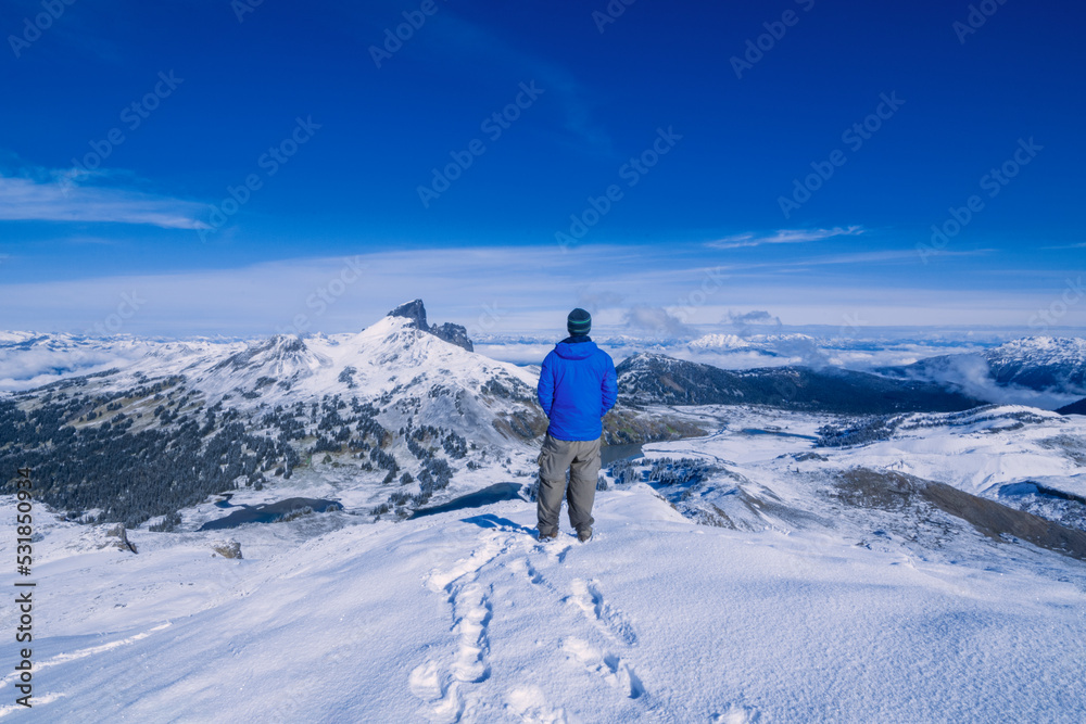 Adventurous athletic male hiker standing in snow at the summit of Panorama Ridge looking at The Black Tusk on a beautiful sunny day.
