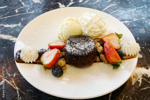 A plate of chocolate cake with vanilla ice cream, berry, and strawberry on a black marble table.