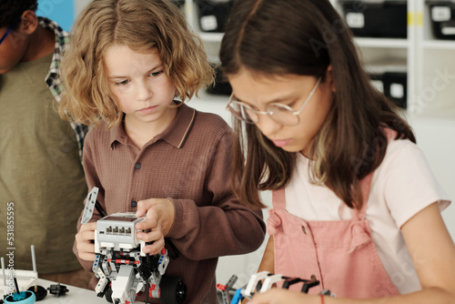 Valokuva Cute sesrious schoolboy looking at his classmate constructing robot while both s