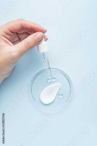 Female hand holding a pipette dropper with face serum or peeling over petri dish with samples of cream and oil. Organic cosmetics. Top view flat lay, copy space. Cosmetics, SPA branding, mock-up.
