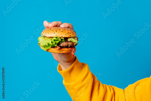 Male hand in yellow clothes hold hamburger on blue background