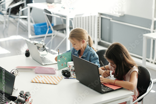 Two clever schoolgirls making notes in their copybooks while sitting in front of laptops by desk and preparing school project