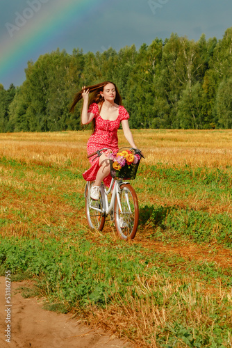 A young beautiful woman rides a bicycle across the field against the backdrop of a forest and a rainbow in the sky and enjoys with the sun