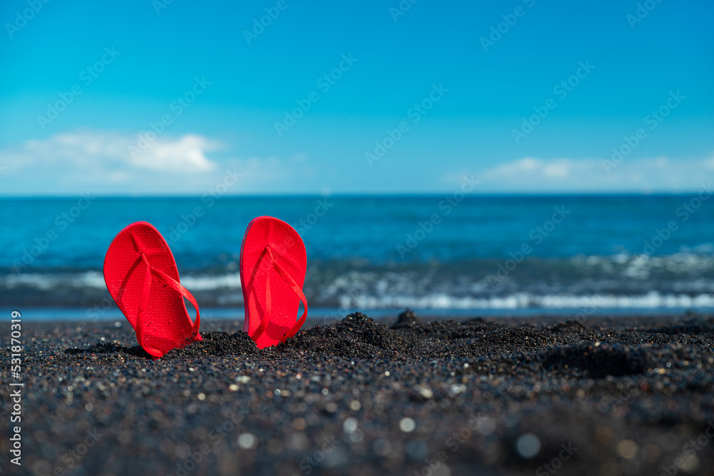Red flip flops on black sandy beach. Calm sparkling ocean and pure blue sky at the background. Summer concept. Red flip flops on black sandy beach. Calm sparkling ocean and pure blue sky at the background. Summer concept.