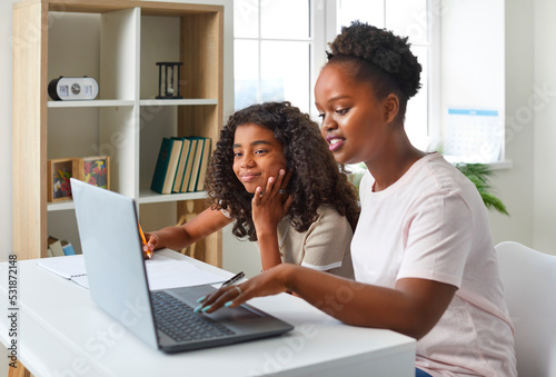 Online education. Caring mother helps her teenage daughter doing homework during remote education. African American woman and dark-skinned girl are using laptop together while sitting at table in room
