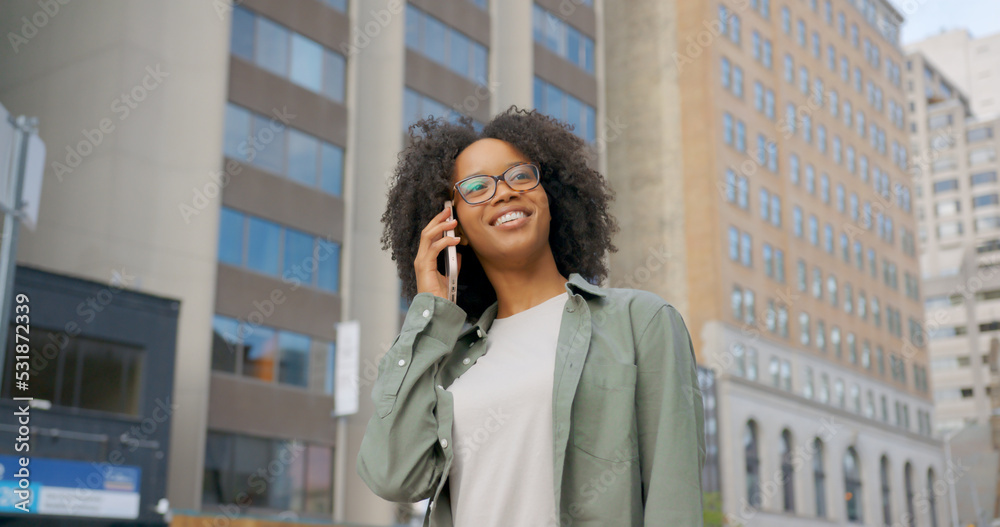 Outdoor shot of positive smiling curly woman having a phone conversation during a walk in modern city, wearing casual green shirt and stylish glasses