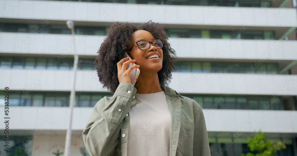 Fototapeta premium Portrait of cheerful mixed-race curly haired woman having a call by her phone outdoors, wearing casual cloth