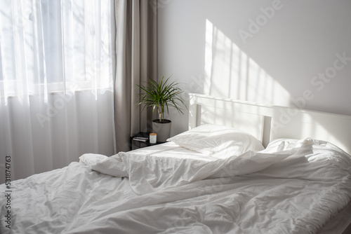 Minimalistic modern bedroom with white bedding and grey curtains. Morning light from the window falling on a bed with white satin bedding.