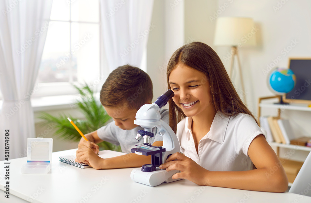 Children and science. Schoolgirl is looking into microscope at home with her younger brother, preparing for science lesson. Funny children sitting at table with microscope and writing in notebook. Children and science. Schoolgirl is looking into microscope at home with her younger brother, preparing for science lesson. Funny children sitting at table with microscope and writing in notebook.