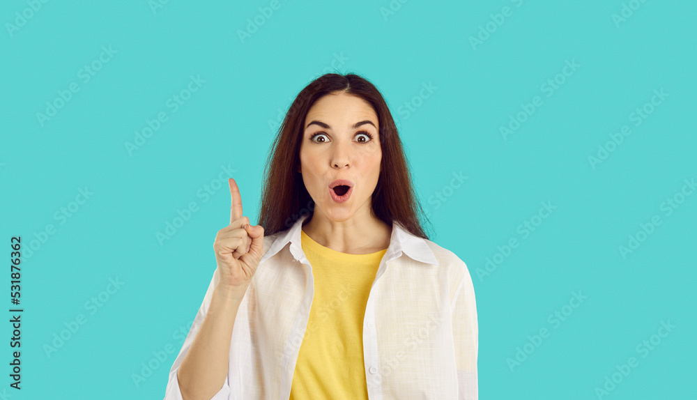 Portrait of emotional funny young woman who came up with great idea or finds solution to problem. Joyful excited girl with wide open mouth raises her finger up on light blue background.