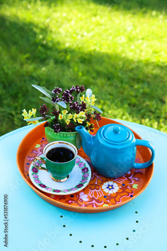 A green coffee cup, a bouquet of wild flowers and a turquoise coffee pot stand on an orange tray. The tray stands on a turquoise table in a sunny garden photo