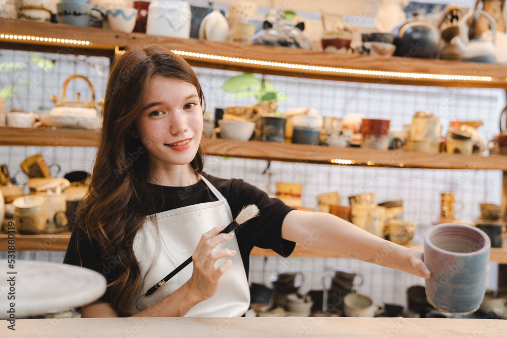 Beautiful young woman holding pottery instrument for scraping, smoothing, shaping and sculpting. Lady siting on bench with pottery wheel and making clay pot