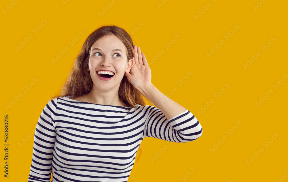 Portrait of intrigued and curious funny young woman eavesdropping someone's interesting conversation. Smiling girl listening with hand to ear while standing near copy space on orange background. Portrait of intrigued and curious funny young woman eavesdropping someone's interesting conversation. Smiling girl listening with hand to ear while standing near copy space on orange background.