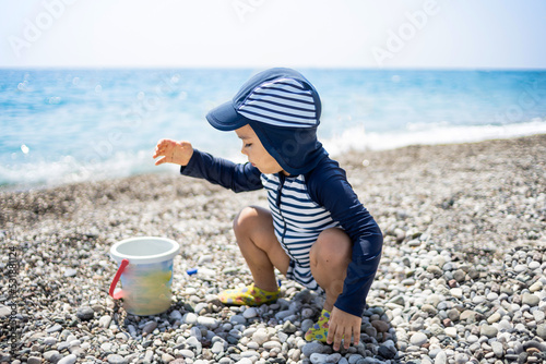 Toddler boy in a swimming suit plays with pebbles on the seashore in a hotel