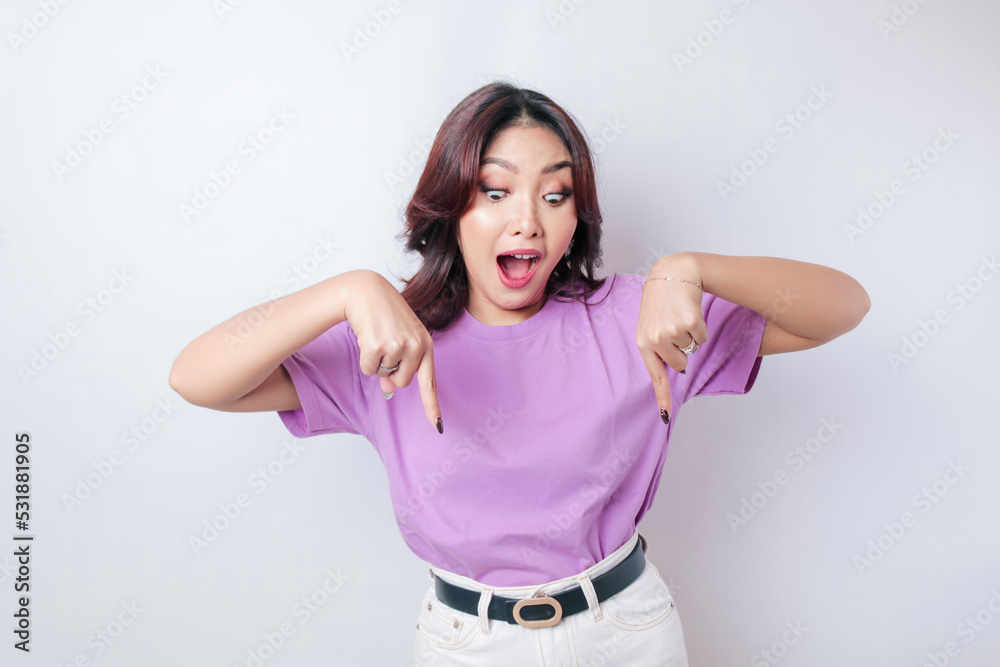 Shocked Asian woman wearing lilac purple t-shirt pointing at the copy space downside her, isolated by white background