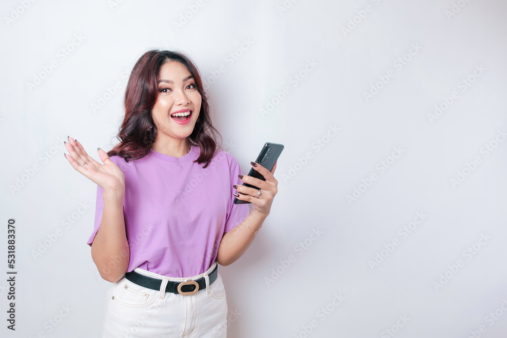 Naklejka premium A portrait of a happy Asian woman is smiling and holding her smartphone wearing a lilac purple t-shirt isolated by a white background