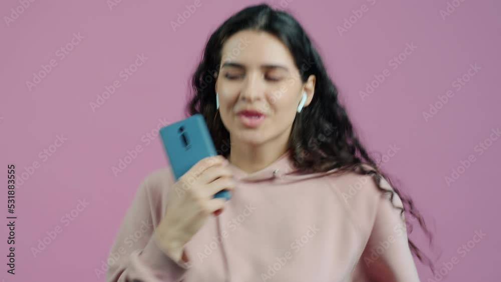 Portrait of carefree young woman dancing using wireless earphones and holding smartphone against pink color background. People and devices concept. Portrait of carefree young woman dancing using wireless earphones and holding smartphone against pink color background. People and devices concept.
