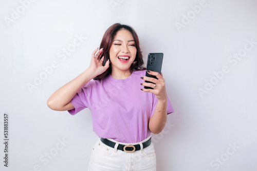 A portrait of a happy Asian woman is smiling and holding her smartphone wearing a lilac purple t-shirt isolated by a white background