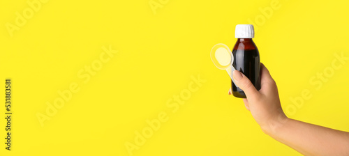 Female hand holding bottle of cough syrup and spoon on yellow background with space for text