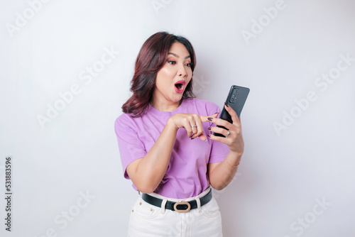 Surprised Asian woman wearing lilac purple t-shirt pointing at her smartphone, isolated by white background