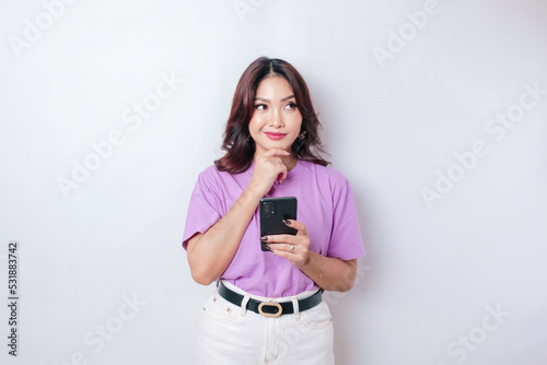 Portrait of a thoughtful young Asian woman wearing lilac purple t-shirt looking aside while holding smartphone