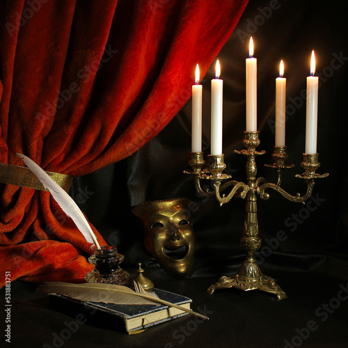  Dramatist's workplace with quill, ink, candelabra, antique book and theatrical mask with dark red curtain. Vintage writing set concept. 