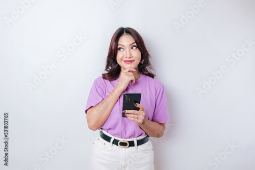 Portrait of a thoughtful young Asian woman wearing lilac purple t-shirt looking aside while holding smartphone