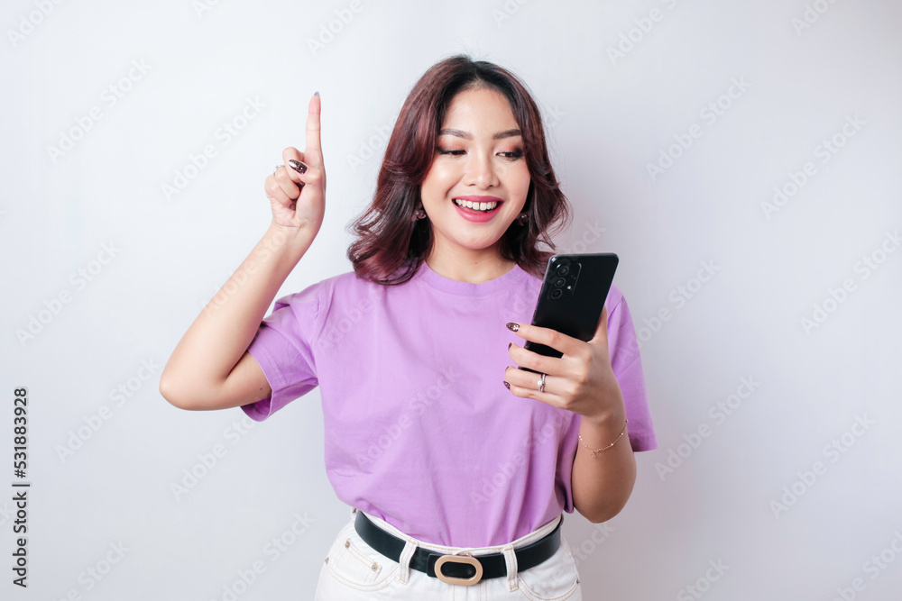 Fototapeta premium Smiling beautiful Asian woman pointing finger upward to empty space while holding her phone isolated white background