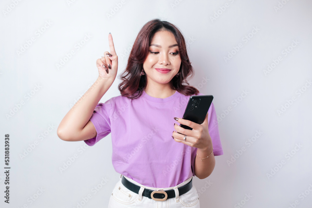 Smiling beautiful Asian woman pointing finger upward to empty space while holding her phone isolated white background