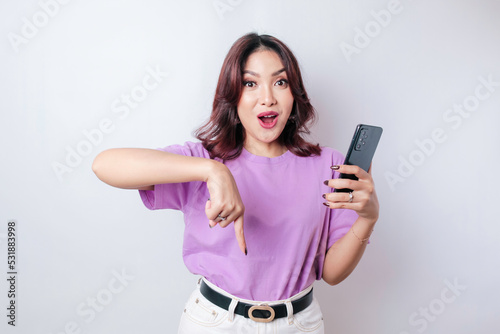 Shocked Asian woman wearing lilac purple shirt pointing at the copy space down below her, isolated by white background