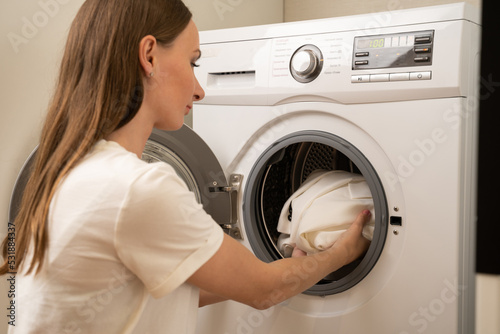 Young friendly woman with long hair, dressed in a white T-shirt, puts dirty white clothes from a basket into a white washing machine in the laundry room