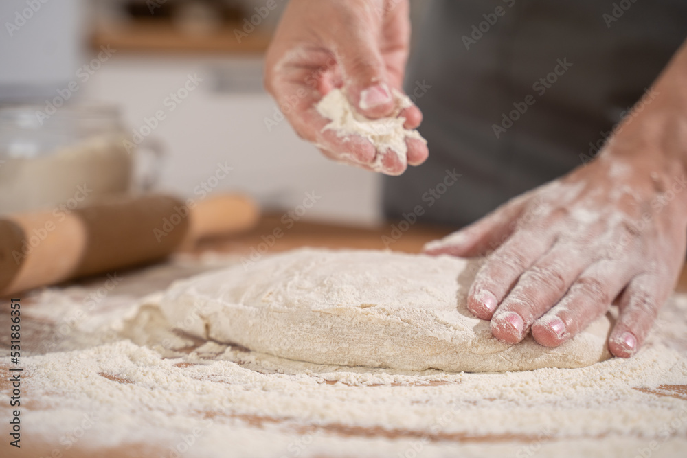 Women's hands pouring flour into raw dough while kneading bread dough on a table full of flour in the bakery kitchen