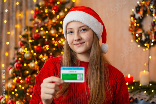 Portrait of a young girl in santa hat holding vaccination certificate in decorated living room. Celebrating Christmas and Coronavirus vaccination concept